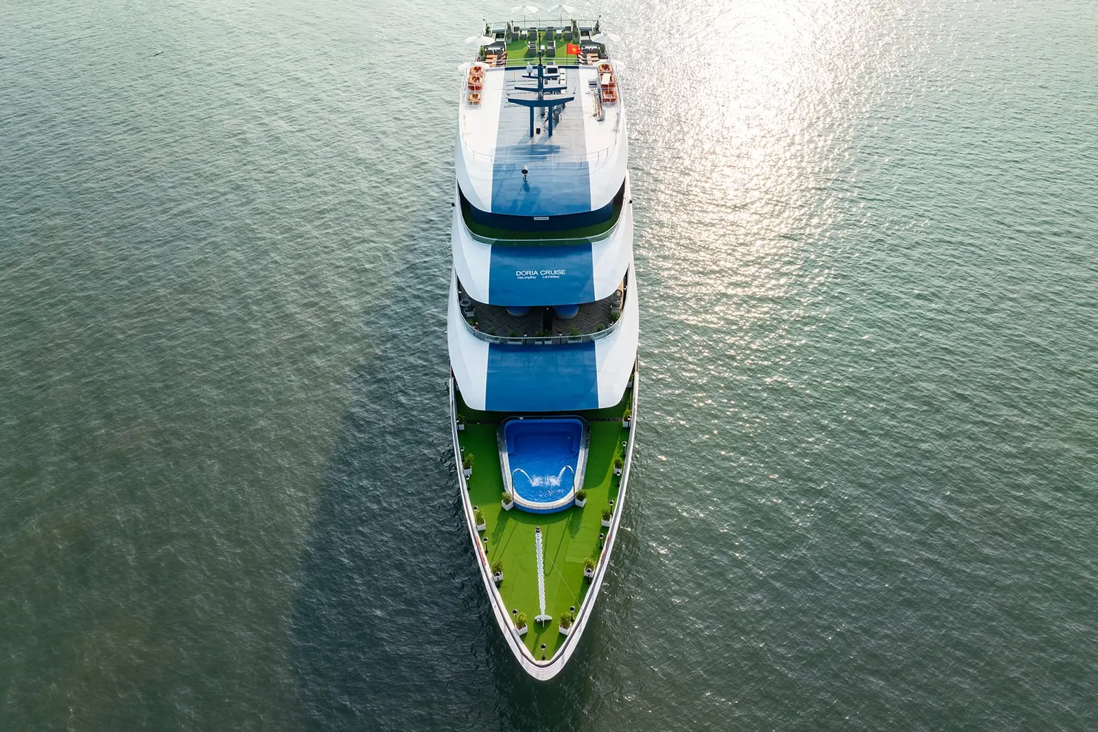 Aerial view of a white multi-deck cruise boat with blue swimming pools and green deck areas moving through calm turquoise water.