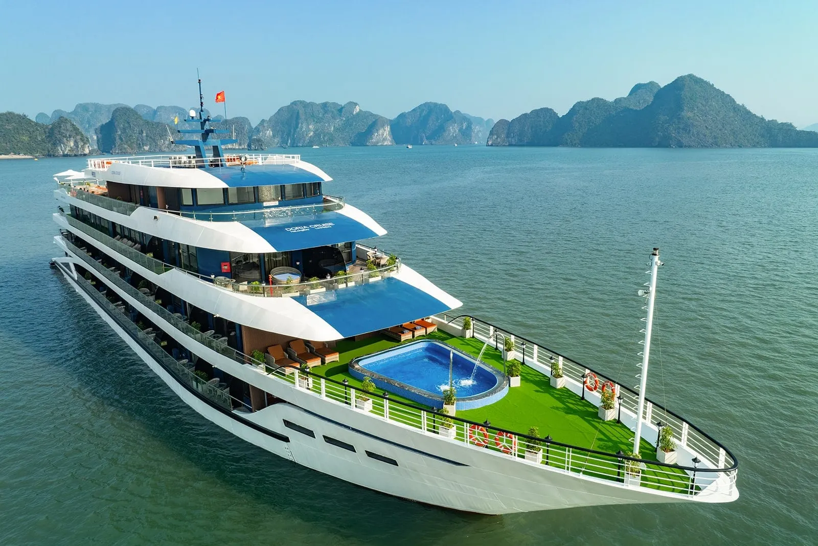 Aerial view of a white multi-deck steel cruise ship with blue canopies, a bow pool and green turf deck, anchored among limestone karst islands under a clear sky.