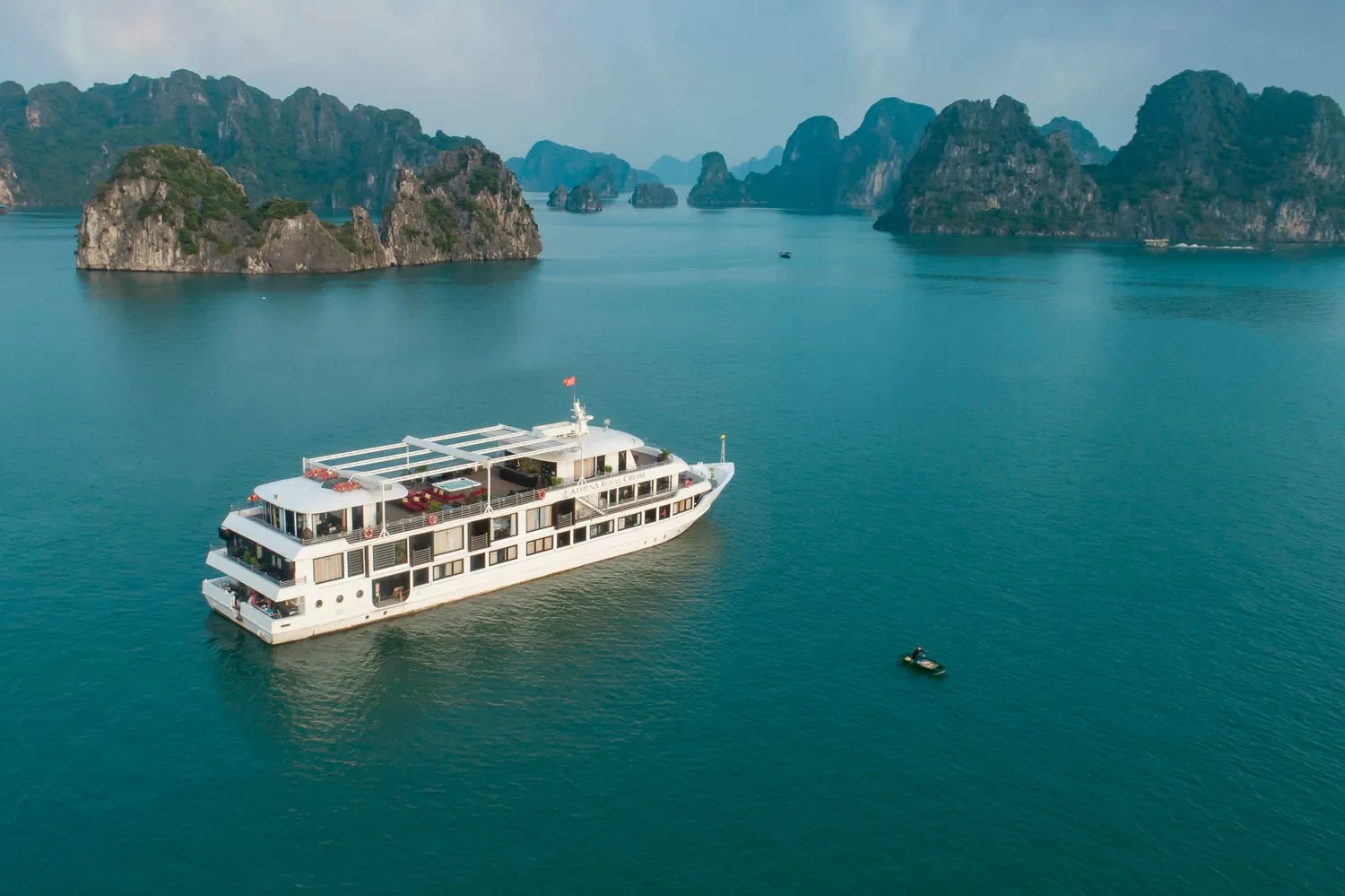 A white multi-deck cruise ship floats in turquoise water surrounded by limestone karst islands under a hazy sky.