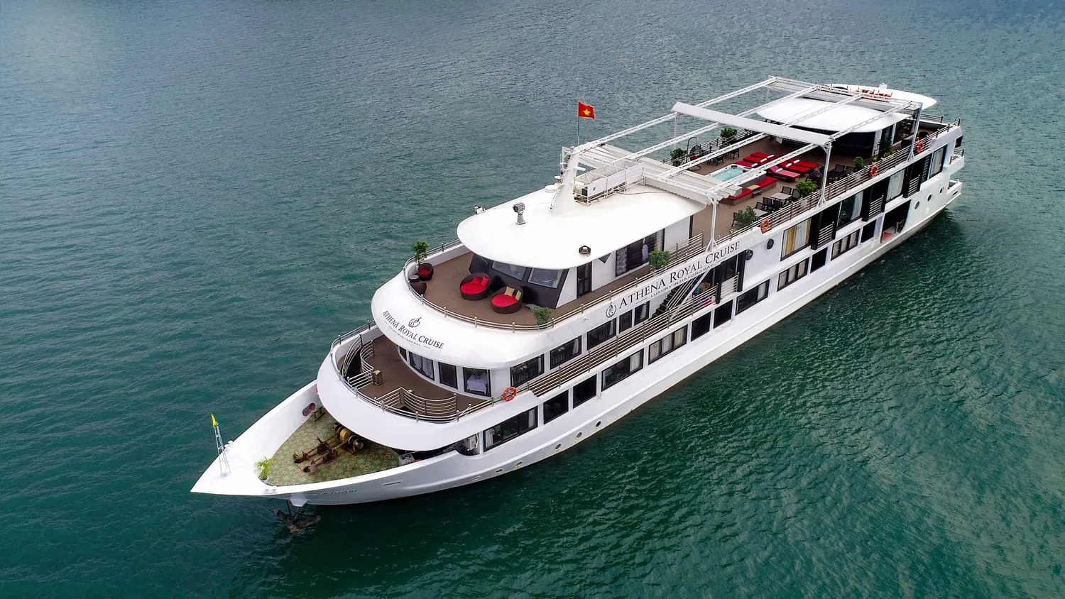 A white multi-deck cruise ship with red cushions on the top deck and the name Athena Royal Cruise painted on its hull floats in calm turquoise water.