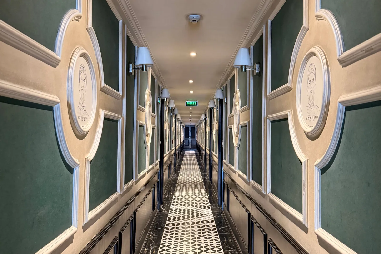 A long symmetrical interior corridor with green panelled walls, white moulding, circular portrait medallions, wall sconces, a black-and-white patterned tile runner and a green exit sign at the far end.