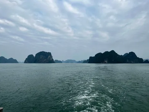 Dark limestone karst islands rise from green-grey water under an overcast sky, with a foamy boat wake in the foreground and a small fixture at the bottom-left corner.