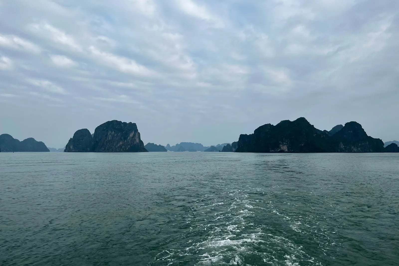Dark limestone karst islands rise from green-grey water under an overcast sky, with a foamy boat wake in the foreground and a small fixture at the bottom-left corner.