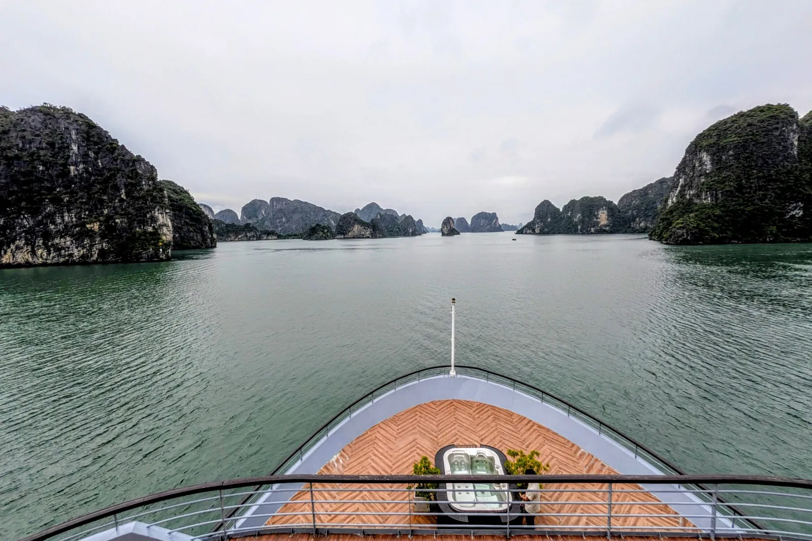 View from an upper deck over the boat's bow with a herringbone wood floor and a small hot tub, looking out across green water toward limestone karst islands under an overcast sky.