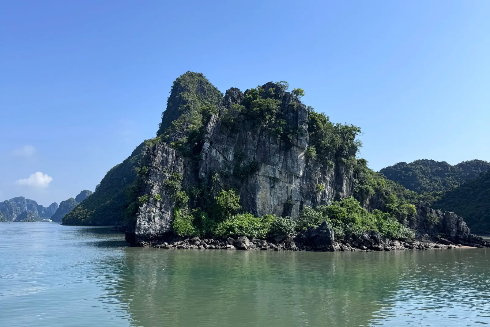 A green-vegetated limestone karst island rises from calm green water under a clear blue sky, with more karst peaks in the distance and a faint wake in the lower left.