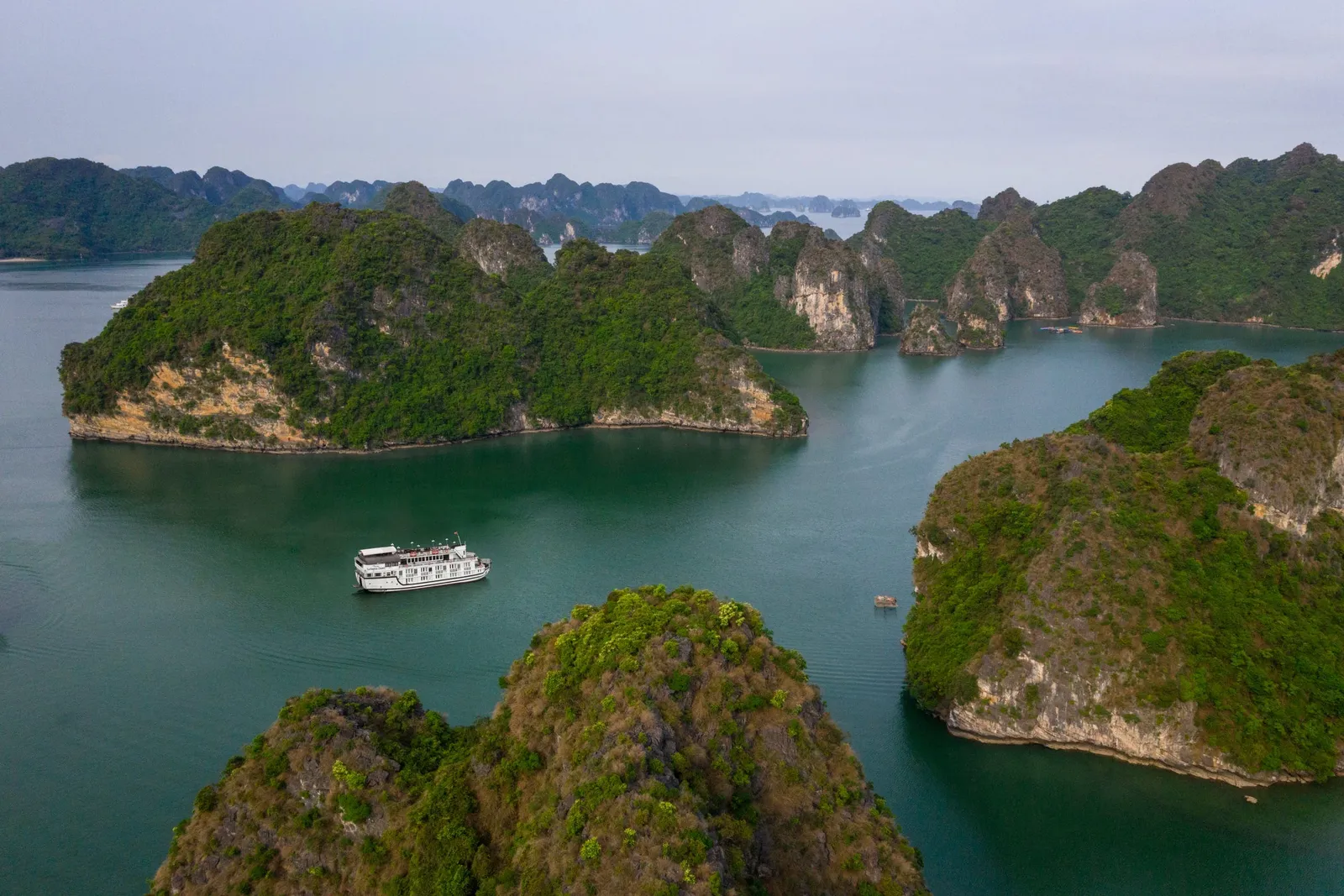 Aerial view of a small white cruise boat on green-teal water surrounded by forested limestone karst islands under a hazy sky.
