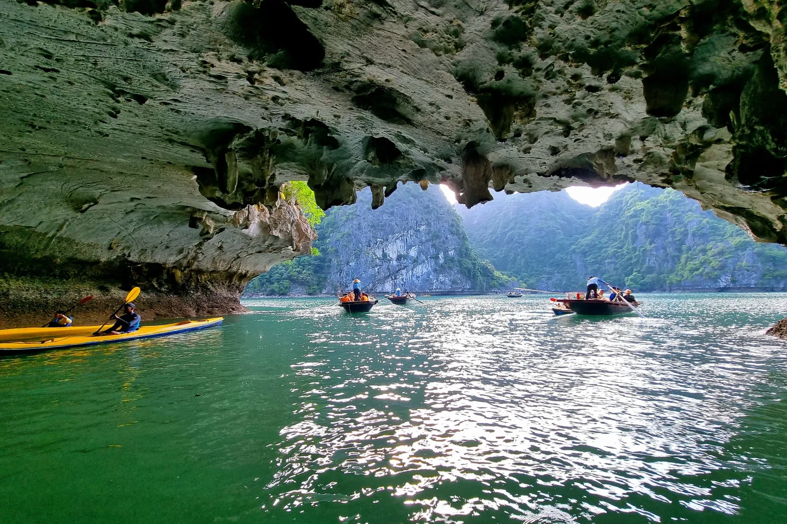 Kayakers and small rowing boats on green water beneath a weathered limestone cave arch, with forested karst cliffs visible beyond the opening.