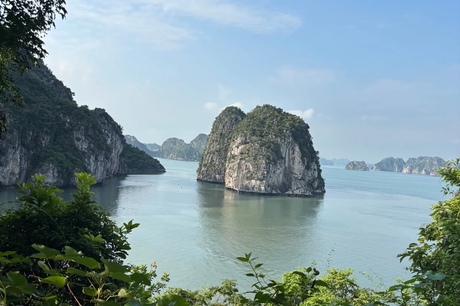 A view through green foliage from a hillside onto calm bay water with a limestone karst outcrop in the center and more karsts fading into the distance under a blue sky.