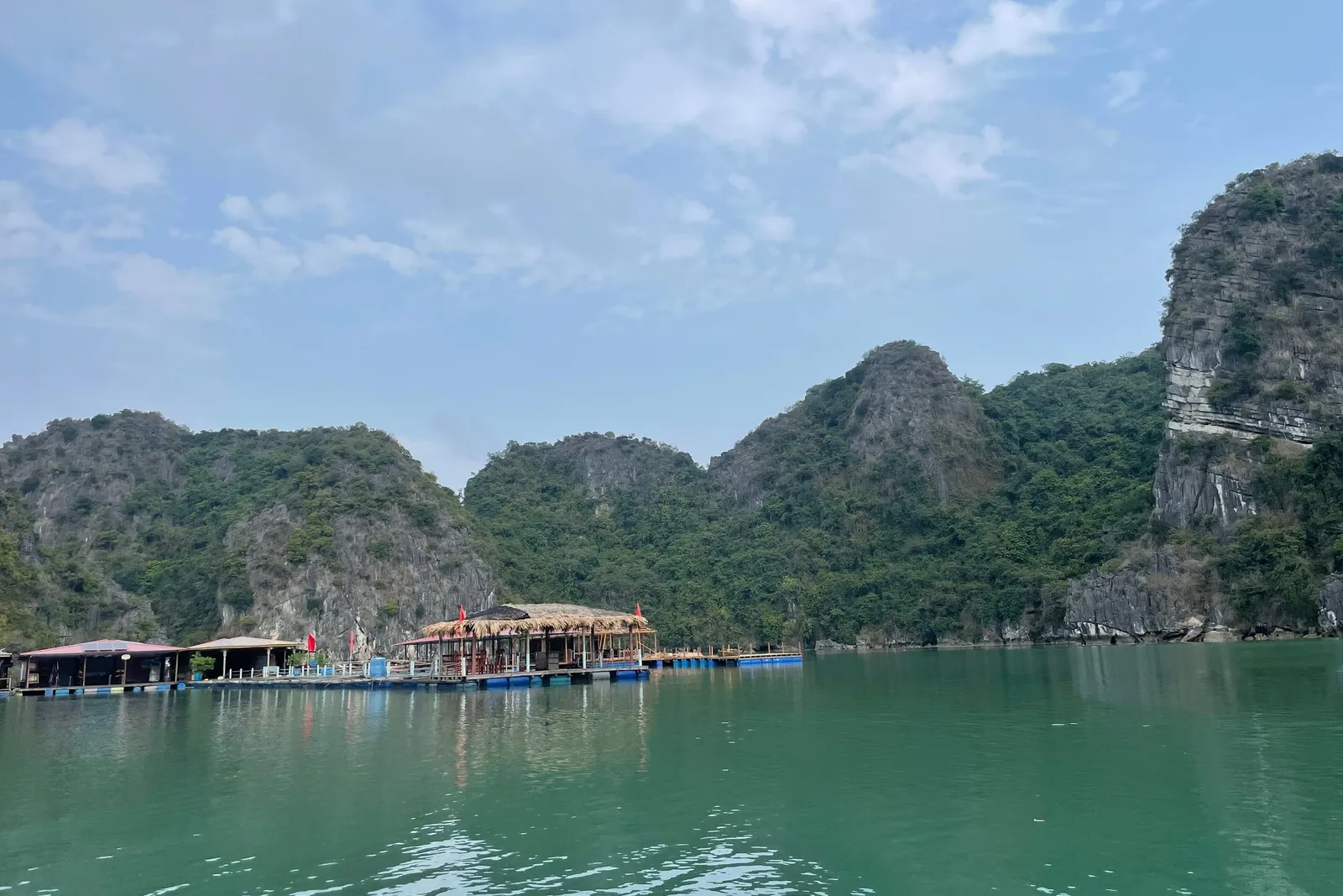 Green water in front of forested limestone karst hills, with a floating platform of thatched-roof and small pavilions with red flags on the left, under a hazy blue sky.