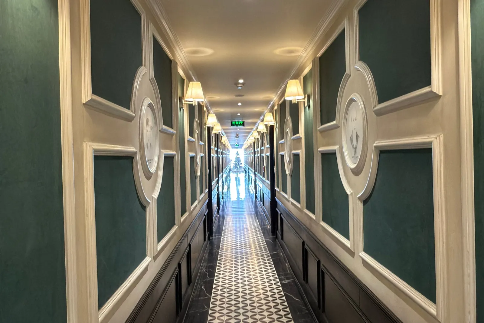A long, narrow corridor with green padded wall panels, cream moldings, round emblem medallions, wall sconces, black marble and patterned tile flooring, and a green EXIT sign at the bright far end.