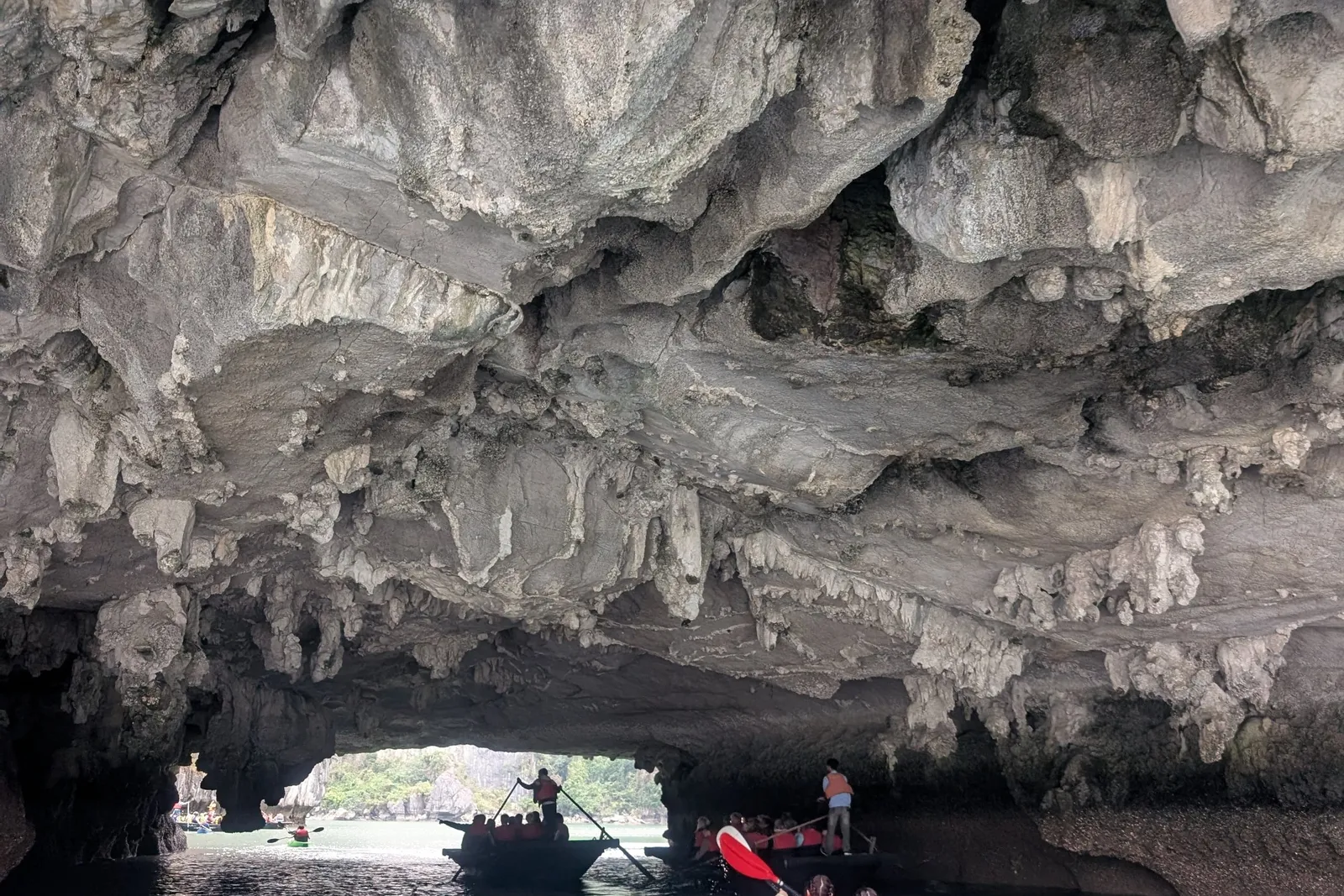 A blue tandem kayak with two people in orange life vests paddles toward a limestone cave opening where rowing boats carrying passengers in life vests wait, with green karst scenery visible beyond.