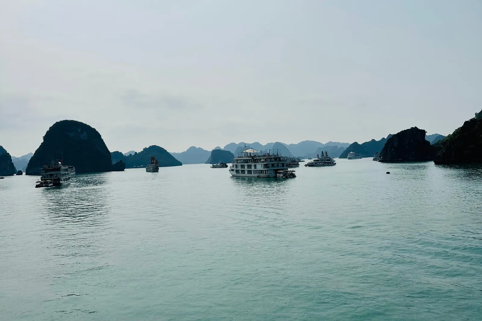 Hazy view across calm turquoise water with several cruise boats anchored among dark limestone karst islands under a pale sky.