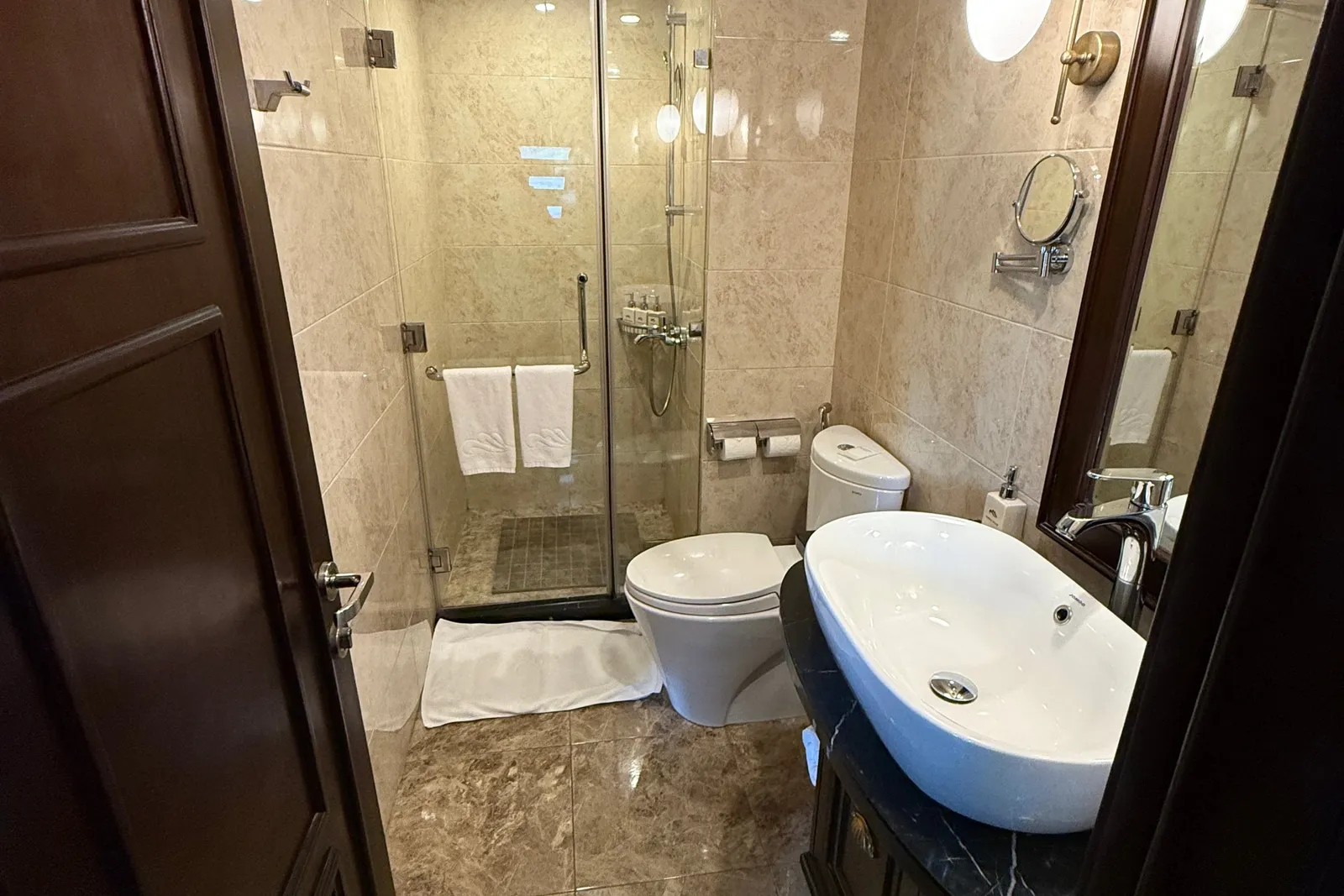 A narrow marble-tiled en-suite bathroom with a glass shower enclosure, white towels, a toilet, a vessel sink on a black marble vanity, a wall mirror and globe wall lights, seen through an open dark wooden door.
