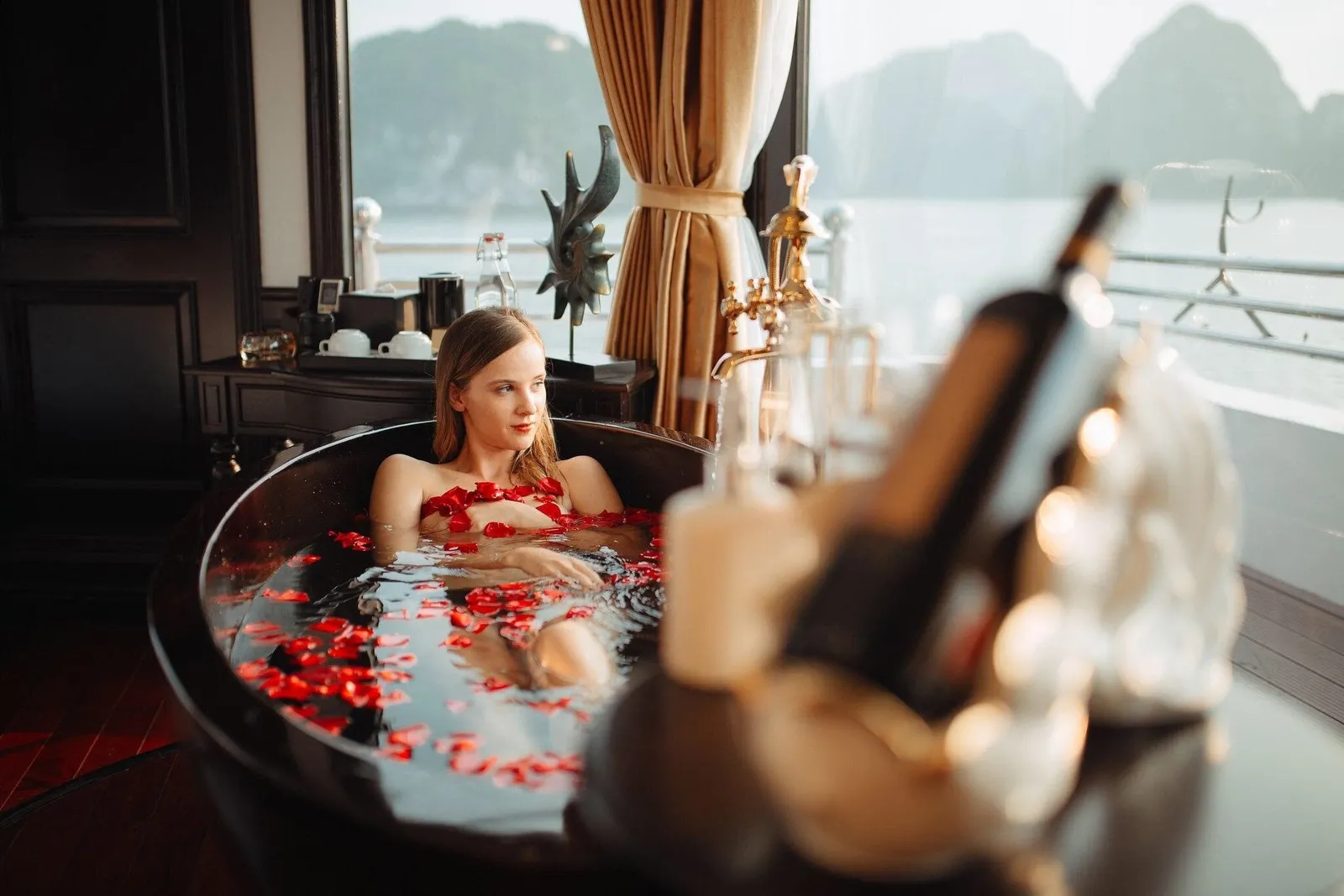 A woman relaxes in a dark round bathtub filled with red rose petals in a cabin with a gold tap, a blurred champagne bottle in the foreground, and a window showing a railing and limestone karst peaks over calm water.