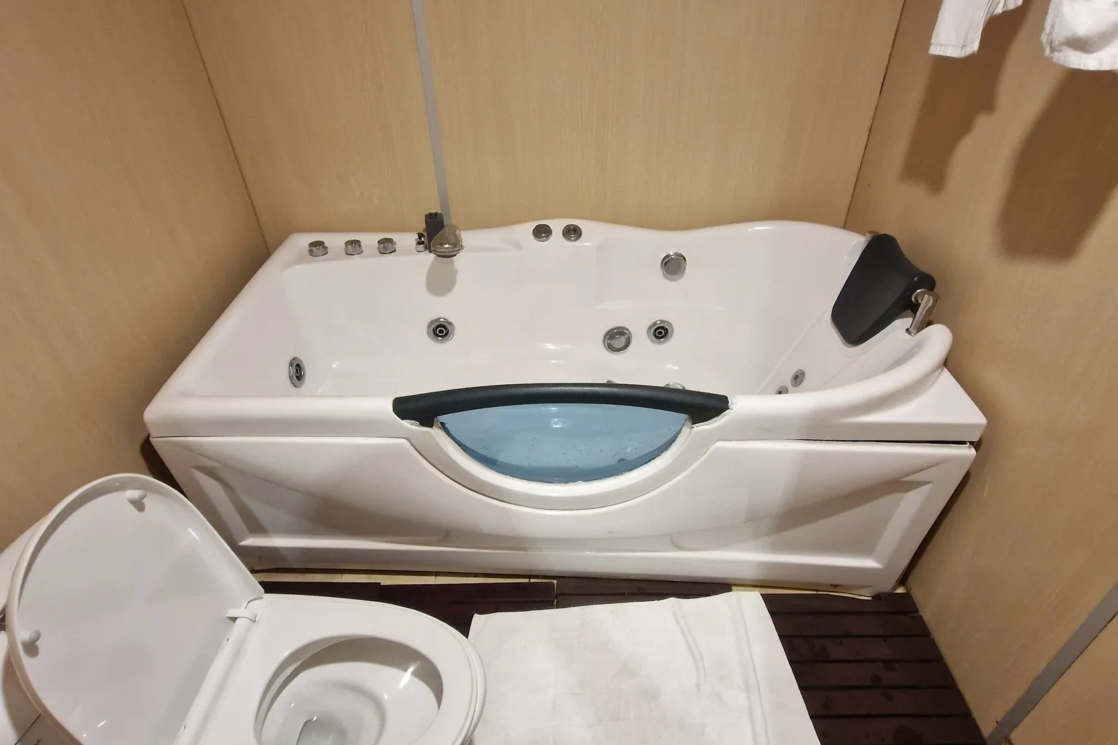 A small en-suite bathroom with a white jacuzzi-style tub, a toilet with its lid up, a white mat on a dark wooden slatted floor, and white towels hanging on a rail, all within beige wood-panelled walls.