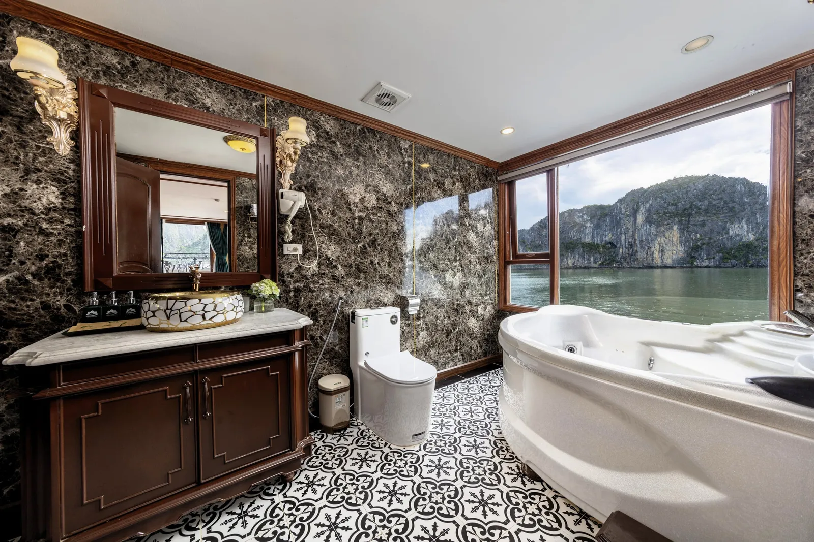A cruise ship bathroom with dark marble walls, wooden vanity, patterned tile floor and a corner jacuzzi tub beside a large window overlooking limestone karst cliffs and calm green water.