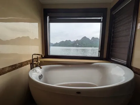 A white freestanding oval bathtub with a brass tap sits under a large window that looks out over calm water toward green limestone karst islands under an overcast sky.