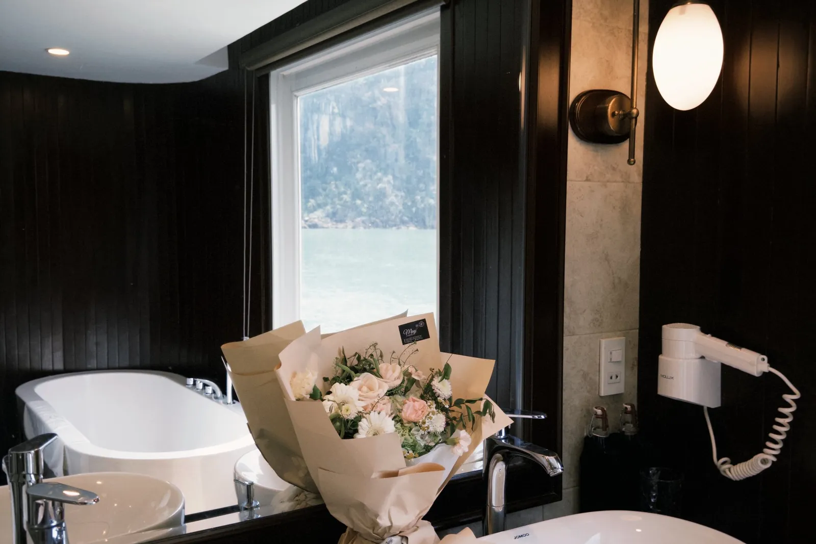 A dark-wood cabin bathroom with two white vessel sinks, a bouquet of flowers wrapped in kraft paper, and a mirror reflecting a bathtub and a window onto green water and a forested limestone cliff.
