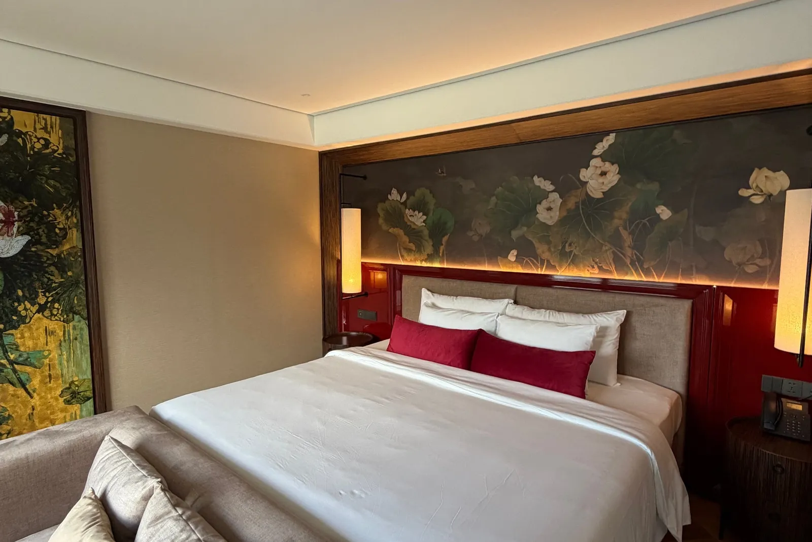 A bedroom with a large white-bedded double bed, red headboard panel, lotus-painted mural, lit wall lamps, a beige sofa with cushions in the foreground and a wood floor, with no visible text, logos or window view.
