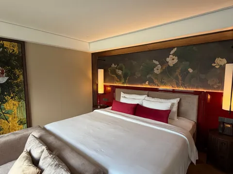A bedroom with a large white-bedded double bed, red headboard panel, lotus-painted mural, lit wall lamps, a beige sofa with cushions in the foreground and a wood floor, with no visible text, logos or window view.