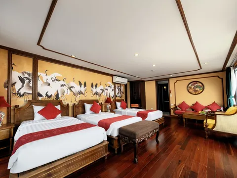 A wood-paneled cabin with three twin beds with white linens and red runners, a painted crane mural on the wall, a sitting area with red cushions, dark hardwood floors, and a window with green curtains.