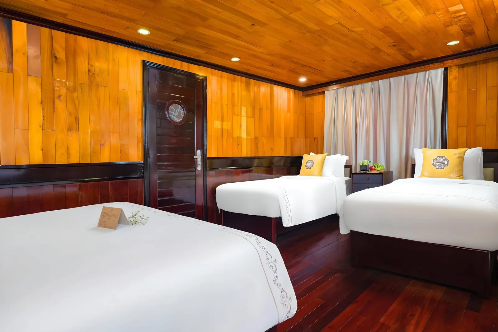 A wood-paneled cabin with three twin beds in white linens, yellow decorative cushions with a medallion emblem, a dark wood door with a round porthole window, cream curtains, and a polished dark hardwood floor.