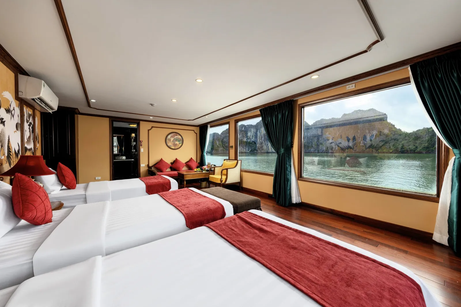 A cruise cabin with three white-linen beds with red runners, a sitting area, and large windows looking out onto calm water and limestone karst cliffs.