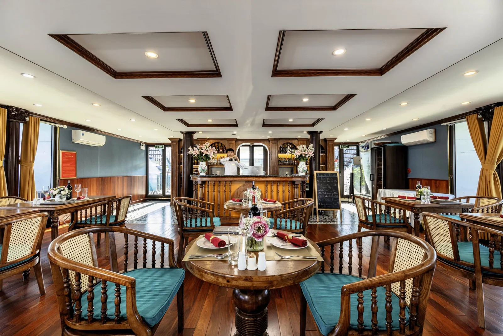 A wood-paneled dining room with set tables, rattan-backed chairs with teal cushions, a bar at the back with a staff member in white, and windows with gold curtains.