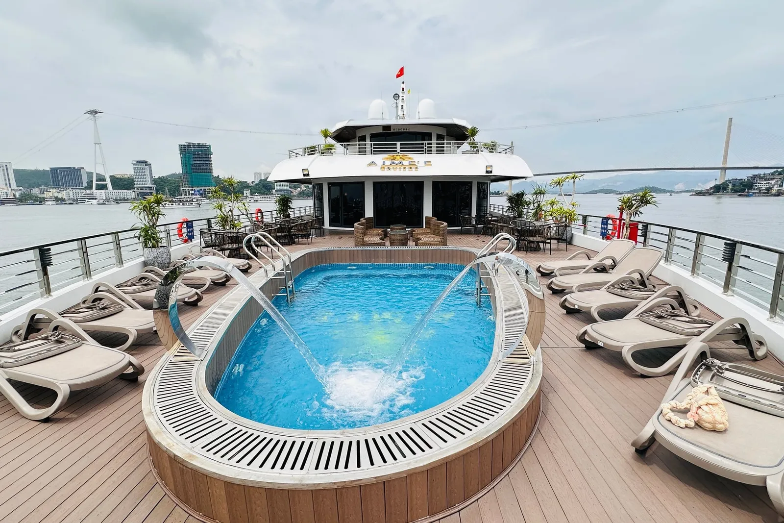 A cruise ship sun deck with an oval plunge pool, loungers and railings, with a branded sign on the upper cabin front and a cable car, bridge and city skyline across the water.