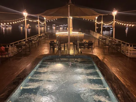 A night view along a boat's upper deck with a bubbling jacuzzi pool in the foreground, tables and chairs, a lit canopy bar, string lights and distant lit boats on dark water.