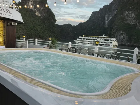A bubbling jacuzzi on a cruise ship's sun deck with string lights and outdoor tables, looking toward limestone karst cliffs and another white cruise ship on the water at dusk, with partial lettering ending in 'UISE' and 'BAY' on the wall of the superstructure.