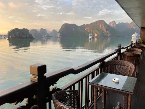 A wooden railing and wicker chairs with a glass-topped table on a boat deck, looking out over calm water toward limestone karst islands and distant cruise boats at sunrise.