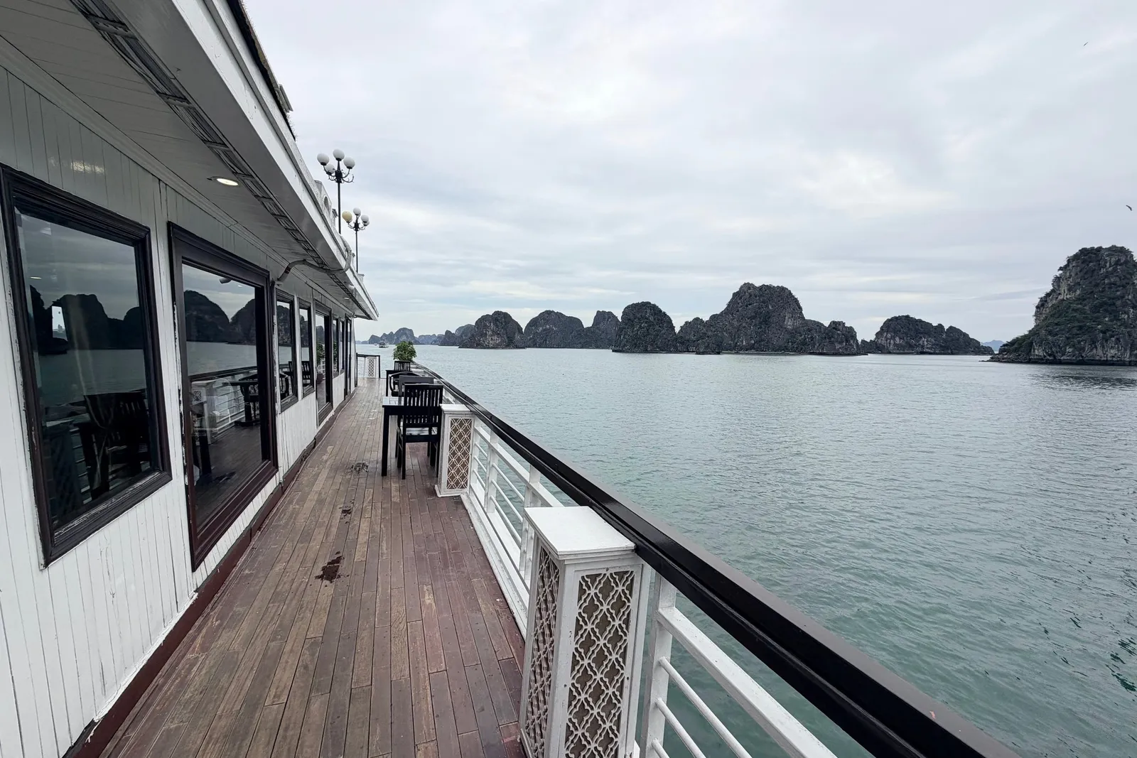 A wooden side deck of a boat with a white railing, dark chairs and a table, and cabin windows, looking out over calm green water toward limestone karst islands under an overcast sky.