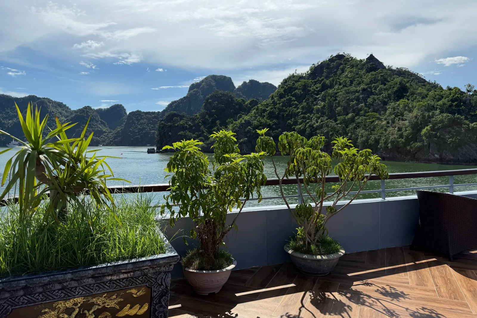 A wooden-floored upper deck with potted plants and a wicker chair, a railing, and green karst islands rising from calm turquoise water under a blue, partly cloudy sky.