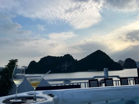 A jacuzzi on a boat's upper deck with a wine glass and a cocktail on its rim, deck chairs and railing behind, and dark limestone islands under a cloudy dusk sky.