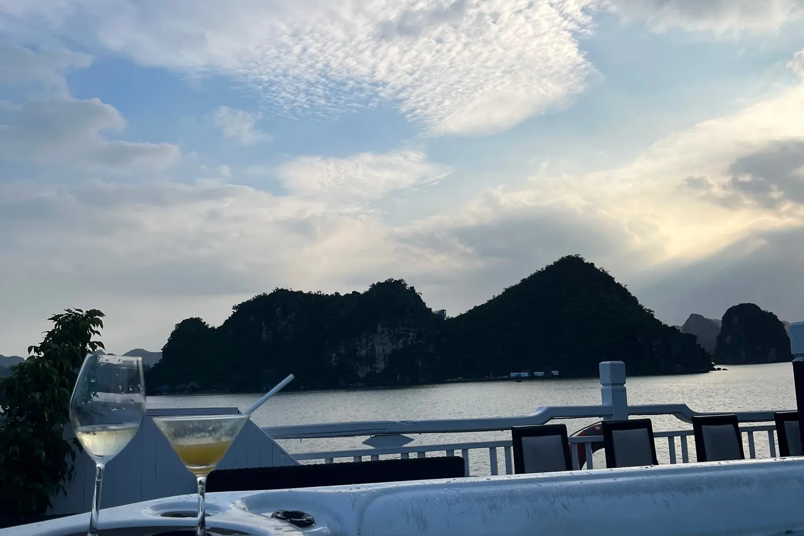 A jacuzzi on a boat's upper deck with a wine glass and a cocktail on its rim, deck chairs and railing behind, and dark limestone islands under a cloudy dusk sky.