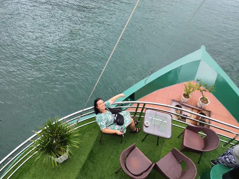 A high-angle view down onto a boat's green bow deck with artificial turf, wicker chairs, a glass table and potted plants, where a smiling woman in a green patterned dress sits waving beside the railing above calm green water.