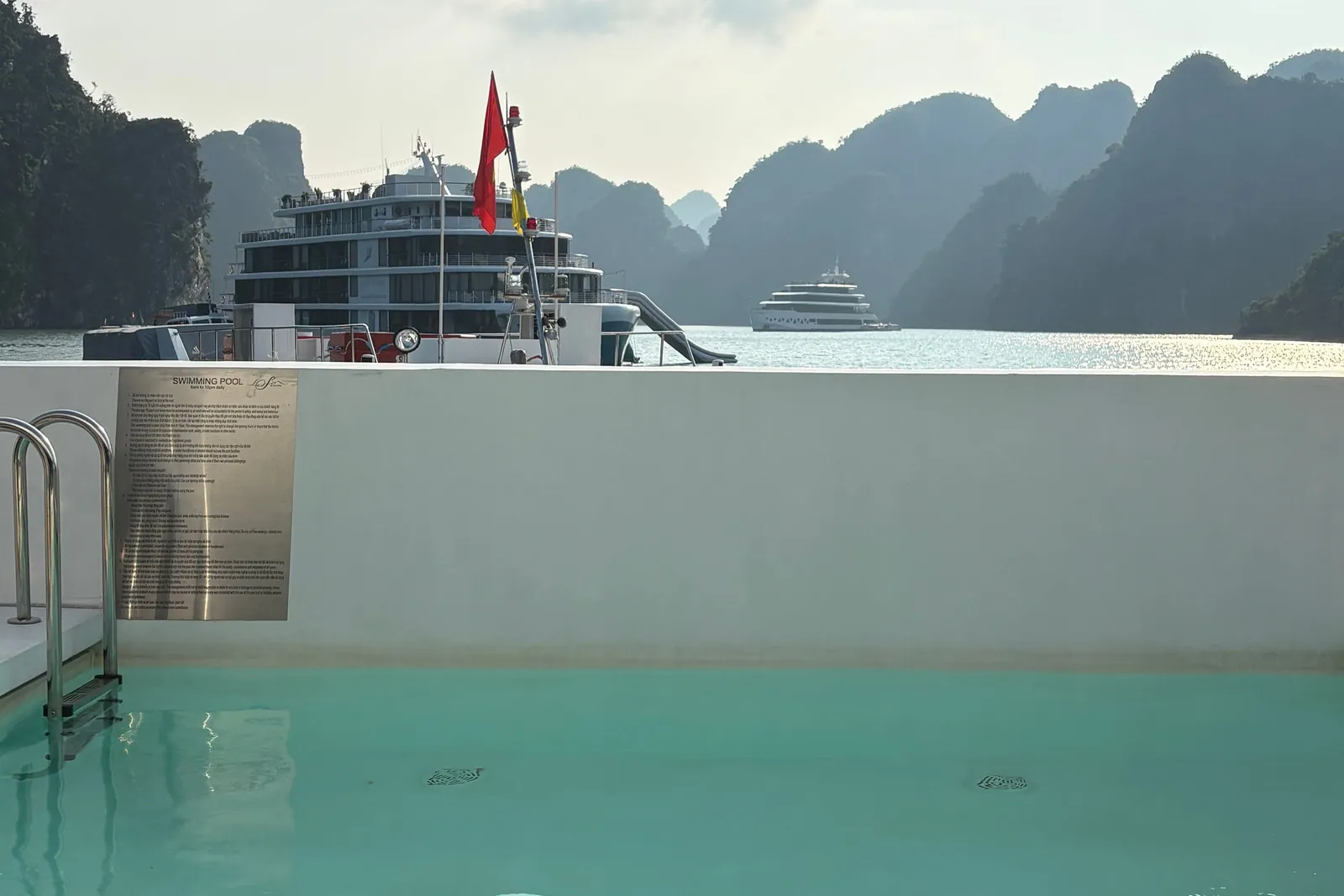A turquoise swimming pool on a boat's upper deck with a metal ladder and a 'Swimming Pool' rules plaque, looking out past the wall to limestone karsts and two other cruise ships on the bay, with a red flag in view.
