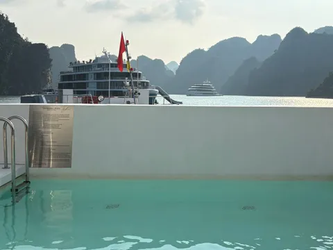 A turquoise swimming pool on a boat's upper deck with a metal ladder and a 'Swimming Pool' rules plaque, looking out past the wall to limestone karsts and two other cruise ships on the bay, with a red flag in view.