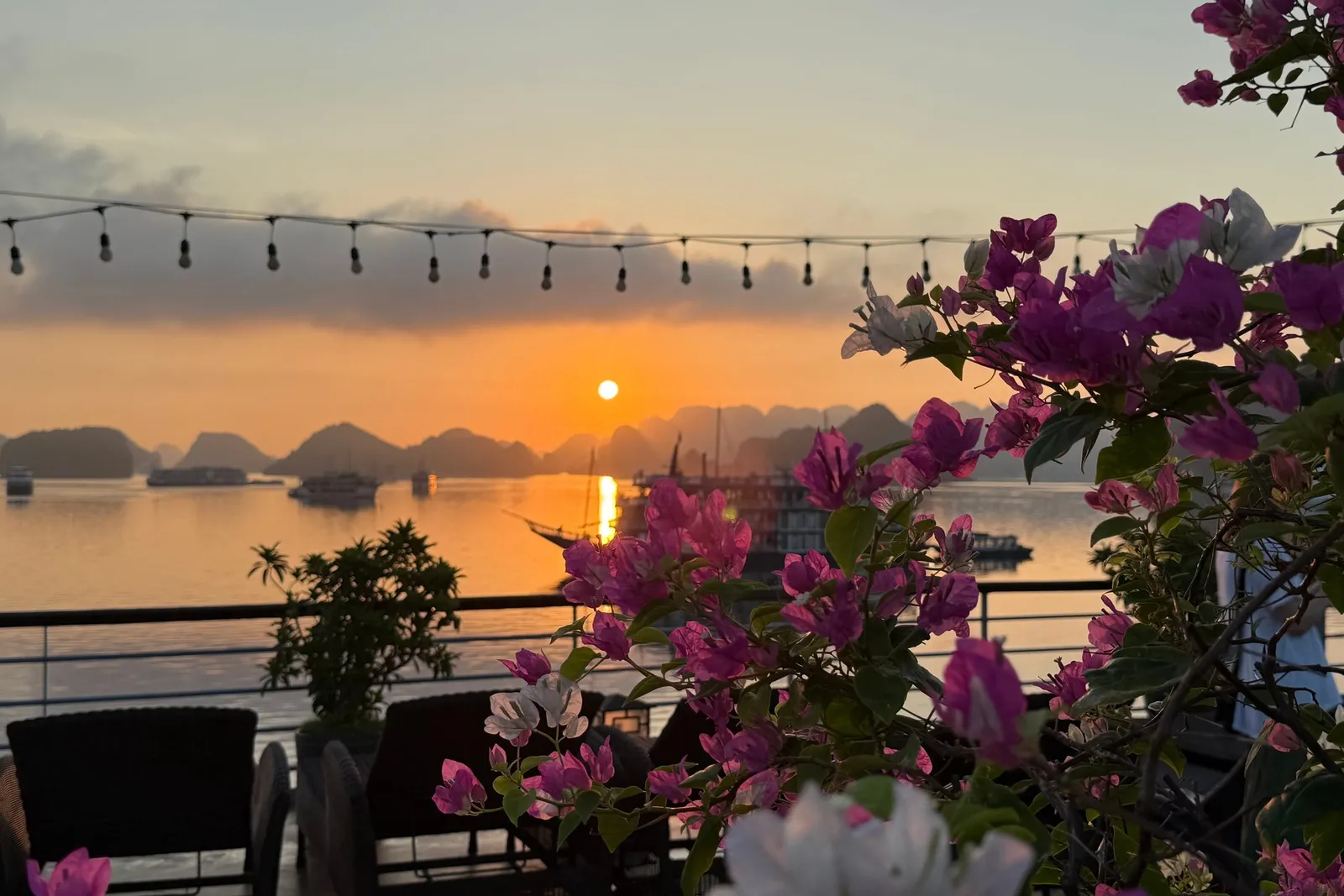 A sunset over a bay with limestone karsts and anchored boats, seen from a railed upper deck with wicker chairs, string lights and pink and white bougainvillea in the foreground.
