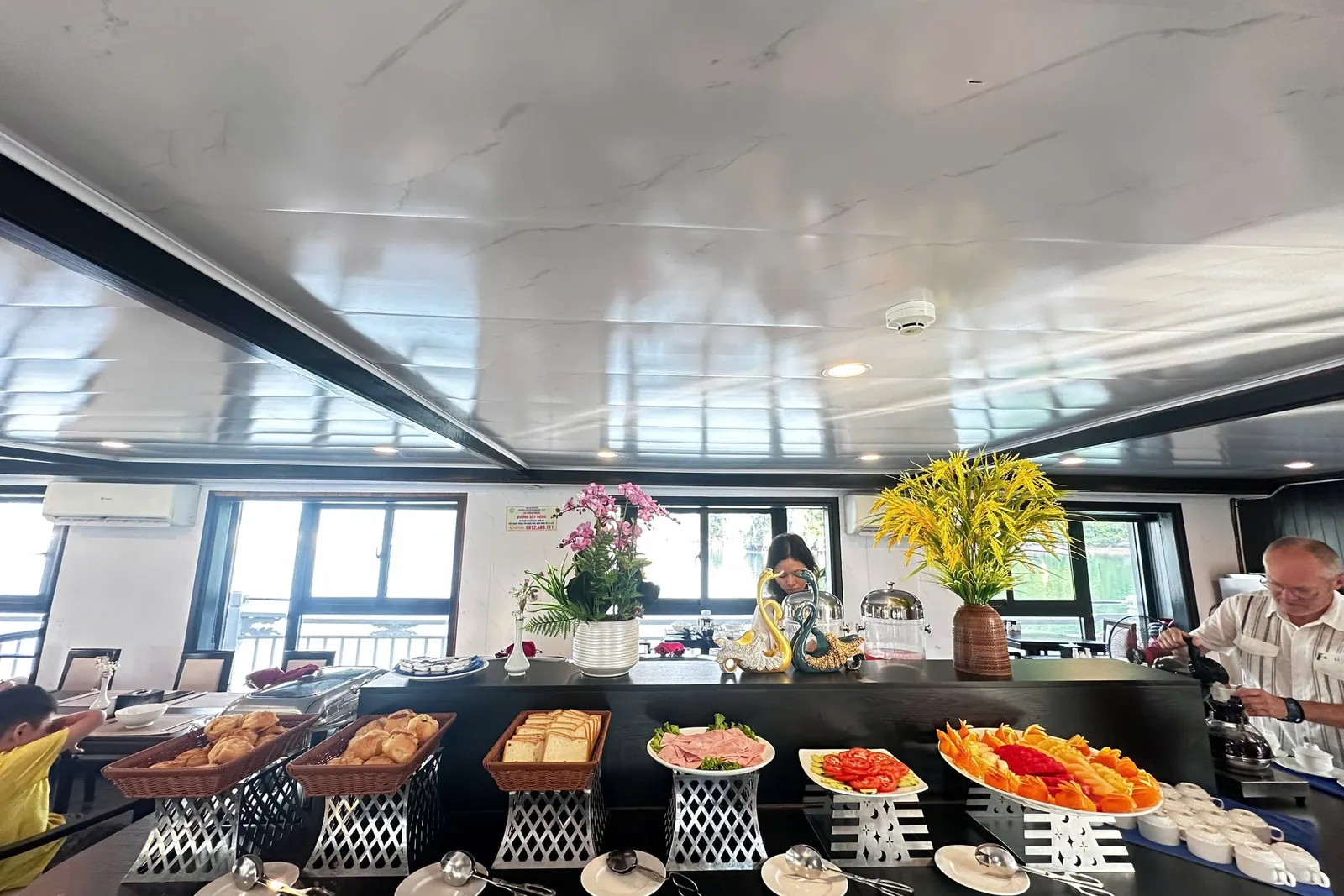 A breakfast buffet counter in an indoor dining room with bread baskets, sliced ham, tomatoes, fruit platter, flower vases and serving tongs, with windows and a railing behind, an older man pouring from a kettle on the right, and a woman standing behind the counter.
