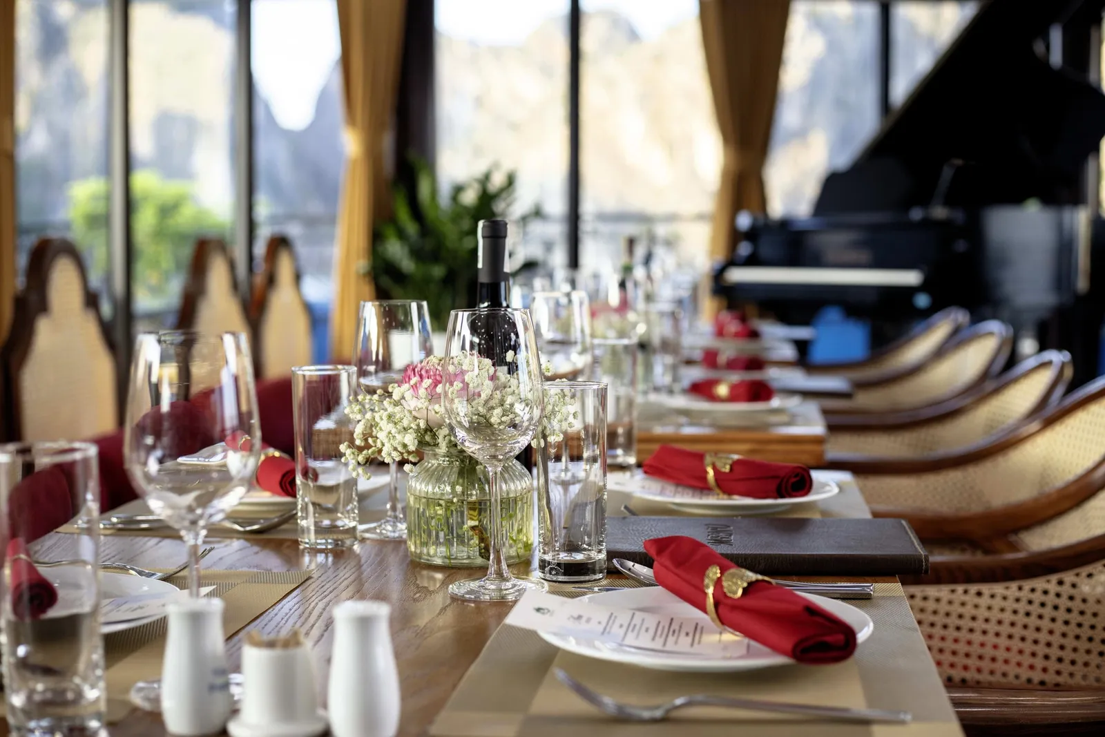 A long dining table set with red napkins, glasses, a flower vase and a menu folder, with rattan chairs, a grand piano and windows showing misty limestone karst hills behind.