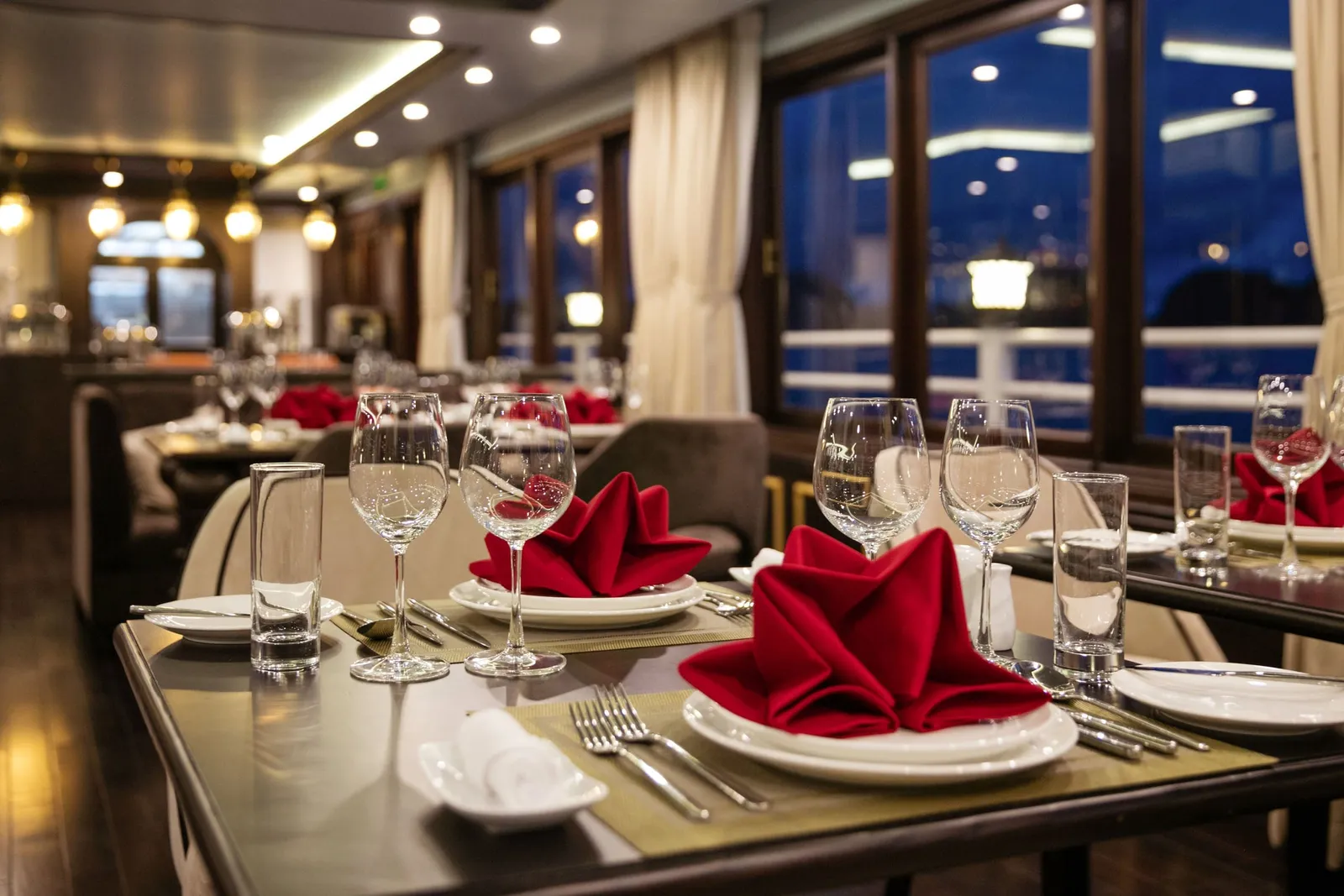 A dining room at night with tables set with red folded napkins, wine glasses, tumblers and cutlery, with large windows showing a railing and dark water outside.