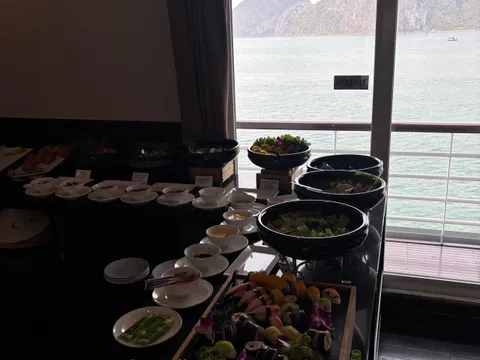 A dimly lit indoor buffet with sushi, salad bowls and small dishes on a black counter, next to a glass door with a 'NO EXIT' sign that looks out over a railing to green water and limestone karst islands.