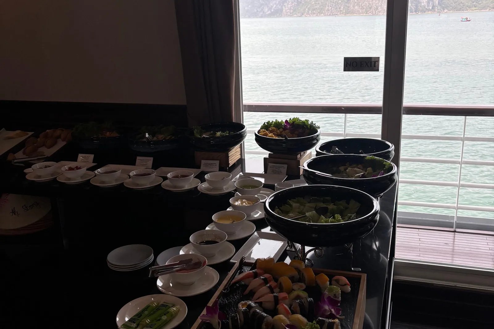 A dimly lit indoor buffet with sushi, salad bowls and small dishes on a black counter, next to a glass door with a 'NO EXIT' sign that looks out over a railing to green water and limestone karst islands.