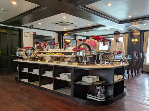 An indoor buffet island with chafing dishes, stacked plates and flower arrangements in a dark-wood dining room, with a waiter in a white shirt and bow tie standing behind it and a window at right.