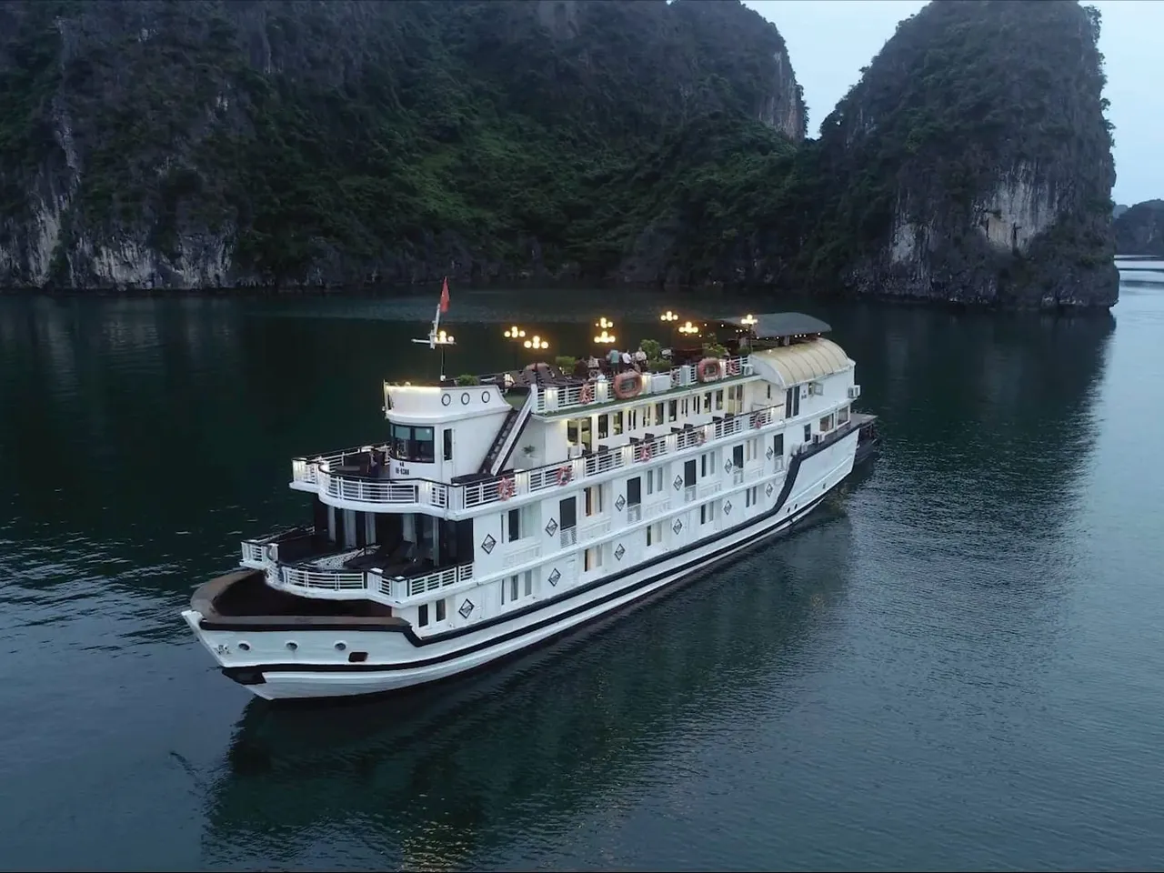 An aerial dusk view of a white multi-deck cruise boat with lit lamps on its upper deck, sailing on calm water beside forested limestone karst cliffs.