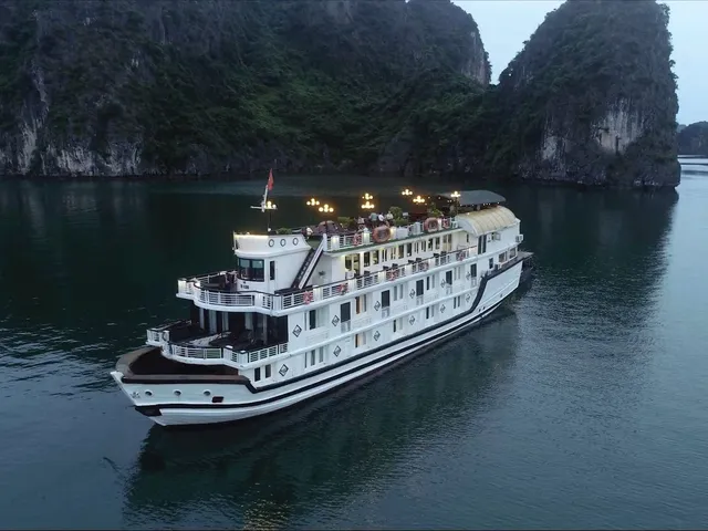 An aerial dusk view of a white multi-deck cruise boat with lit lamps on its upper deck, sailing on calm water beside forested limestone karst cliffs.