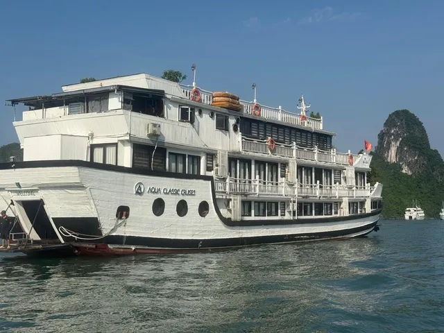 A white multi-deck cruise boat with 'Aqua Classic Cruises' and a logo painted on its hull sits on green water near a forested limestone karst, with a Vietnamese flag at the stern, a person standing on the rear platform, and another boat in the distance, photographed from another vessel on the water.