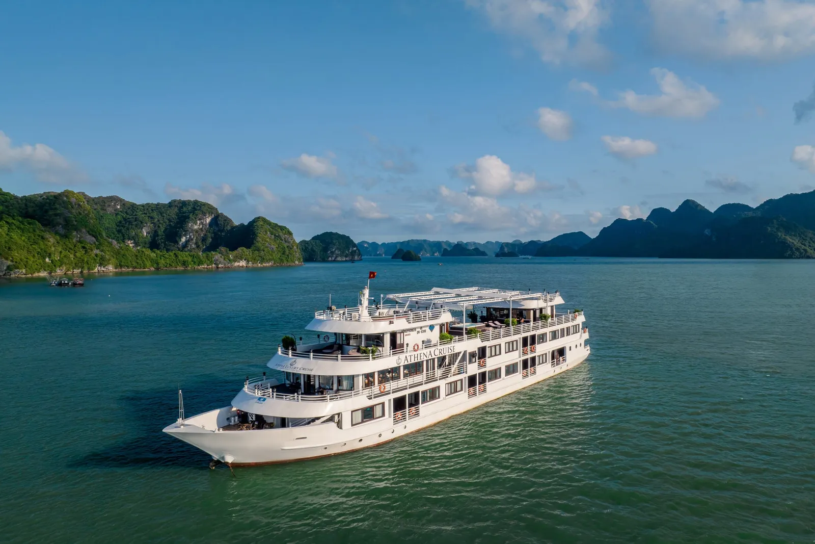 Aerial view of a white multi-deck cruise ship with 'Athena Cruise' lettering on its side and a Vietnamese flag, sailing on green water among forested limestone karst islands under a blue sky.